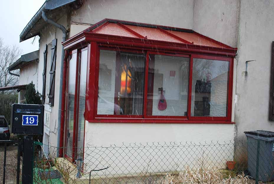 Petit porche fermé rouge et blanc avec une fenêtre éclairée sur une maison, à côté d'un portail bleu portant le numéro 19