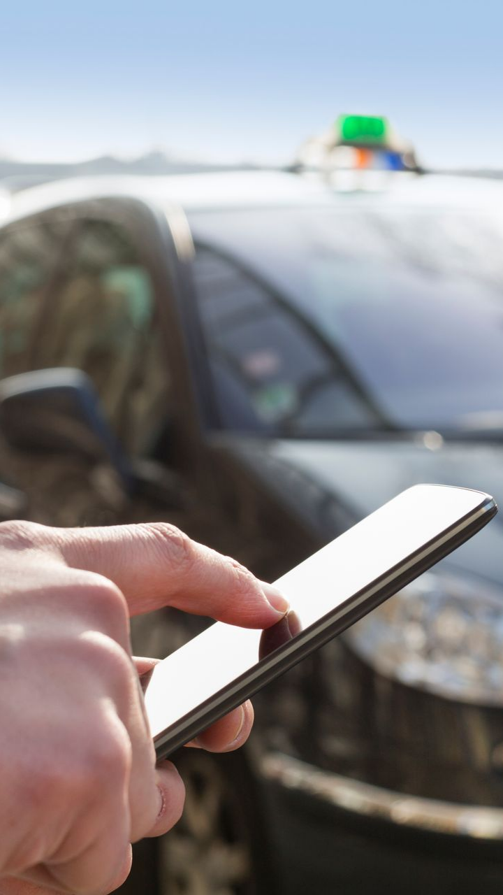 Une personne utilise un téléphone pour appeler un taxi. La voiture est noire, avec un lumignon de taxi en arrière-plan.