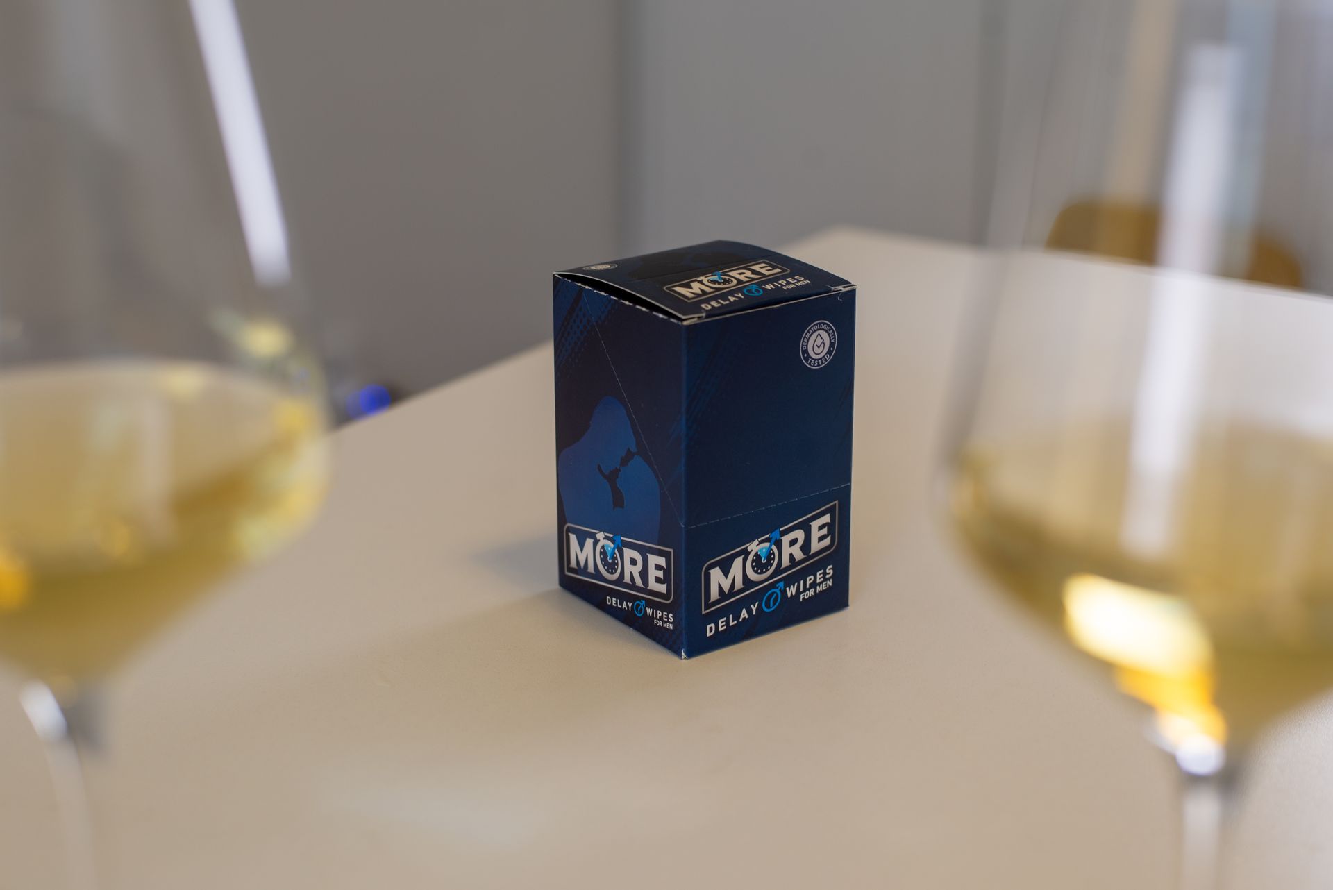 Eine blaue Schachtel mit einem Produkt der Marke „MORE“ steht auf einem weissen Tisch zwischen zwei Gläsern Weisswein.