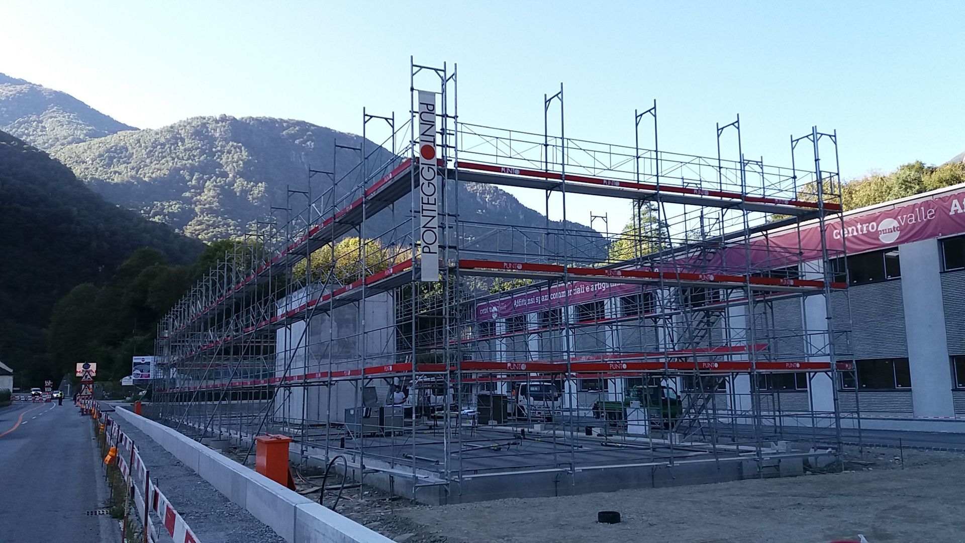 Cantiere con impalcature davanti a un edificio; montagne sullo sfondo.