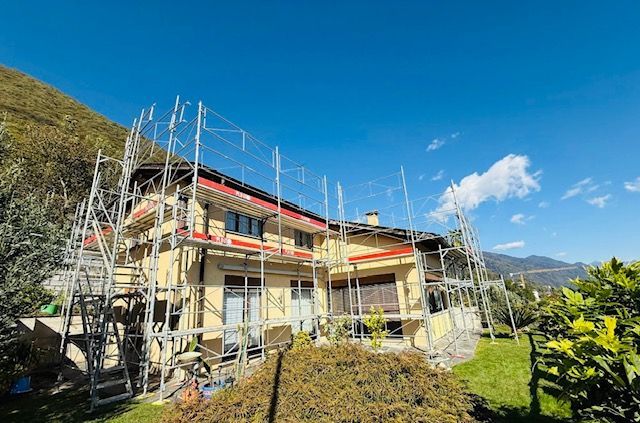 Casa in ristrutturazione, impalcature, muri gialli, prato verde, montagne sullo sfondo, cielo azzurro.