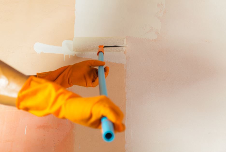 Manos con guantes naranjas pintando una pared de blanco con un rodillo.