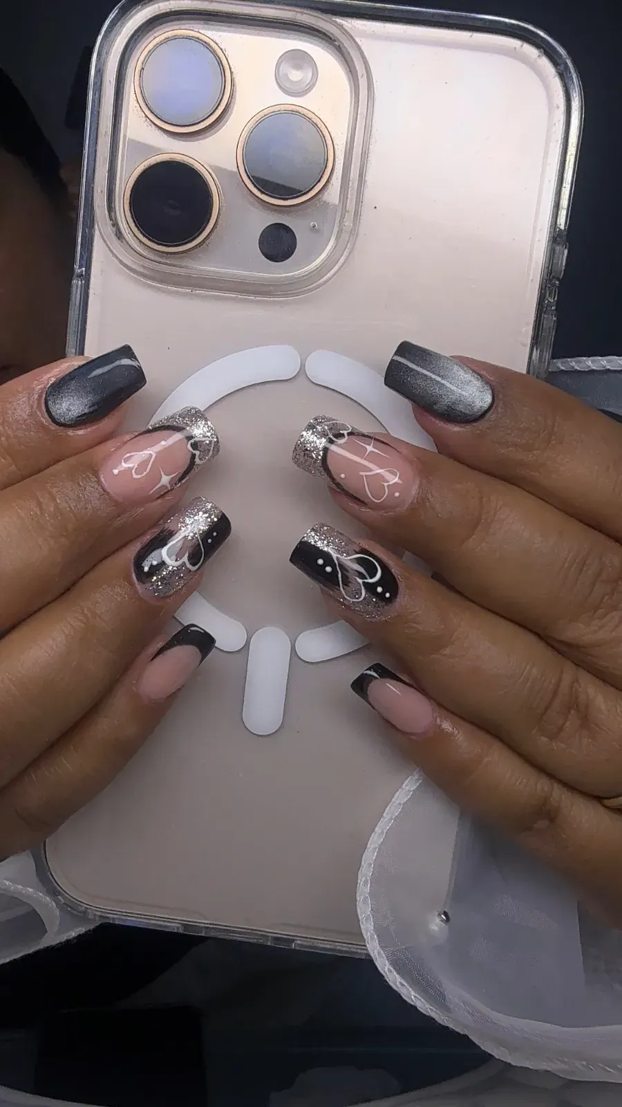 Manos con uñas decoradas con motivos metálicos sujetando una funda transparente para teléfono con un anillo MagSafe blanco.