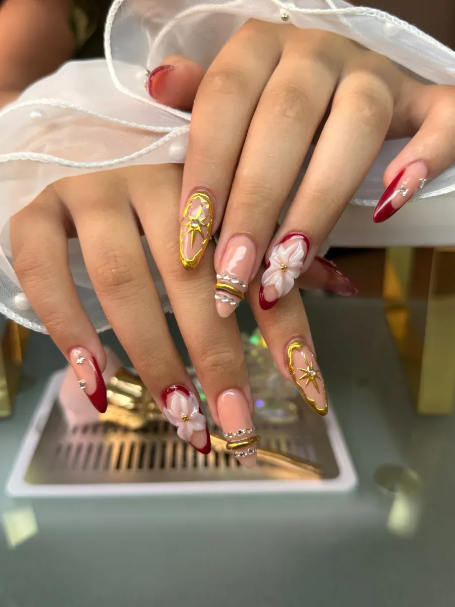 Primer plano de uñas acrílicas largas y decoradas con diseños dorados y rojos, sostenidas sobre un dispositivo blanco.