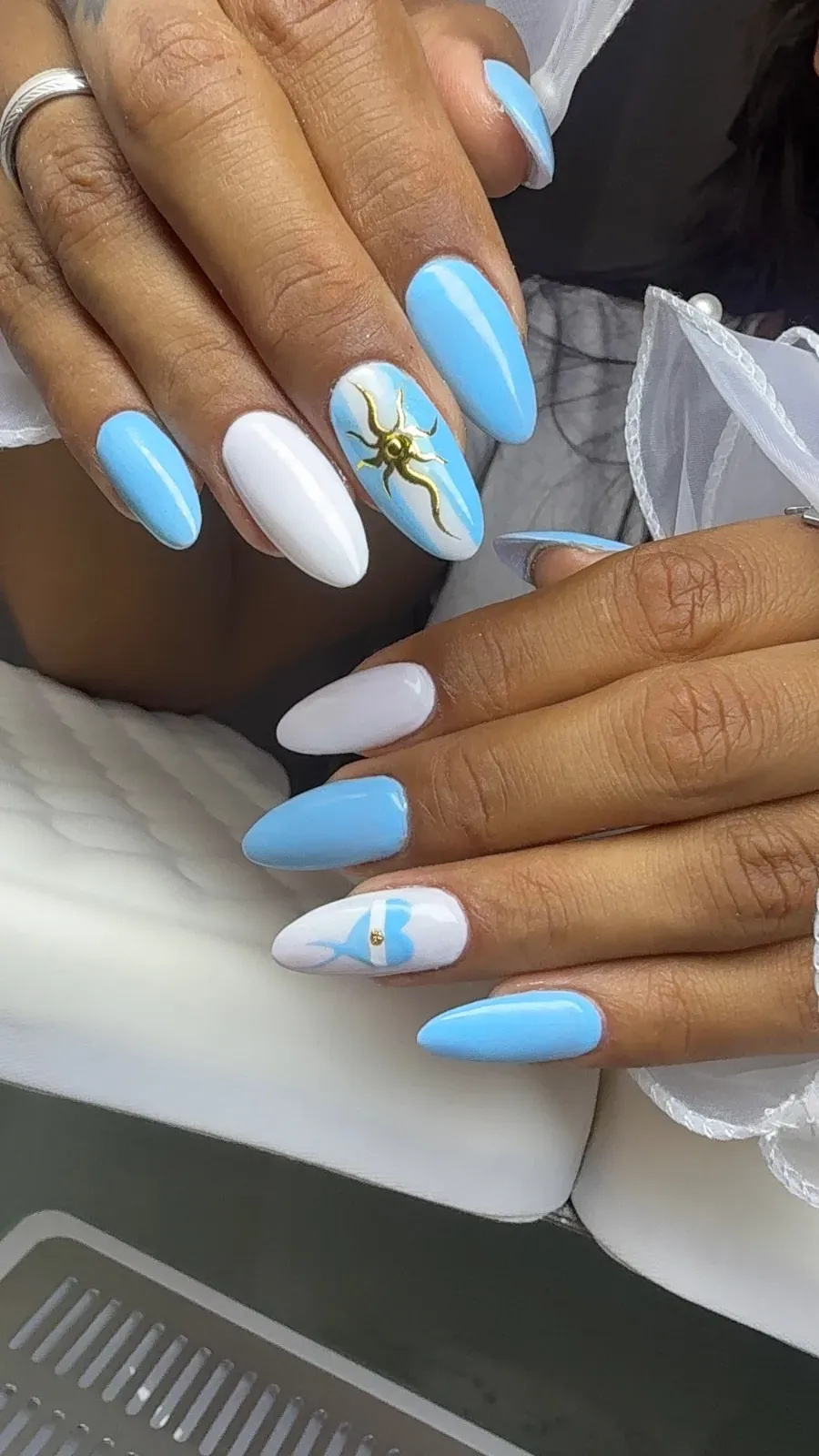 Manos con uñas largas de color azul pastel y blanco, algunas con diseños florales, descansando sobre una silla.