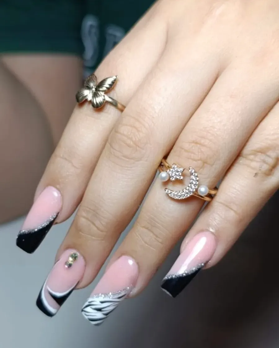 Mano con manicura francesa rosa y negra y anillos dorados sobre un fondo desenfocado.