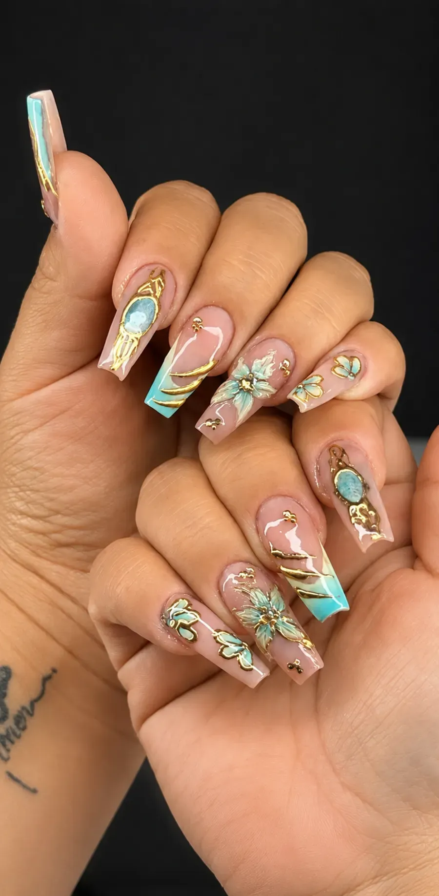 Mano con uñas acrílicas largas iridiscentes en tonos pastel y detalles florales dorados sobre un fondo oscuro.