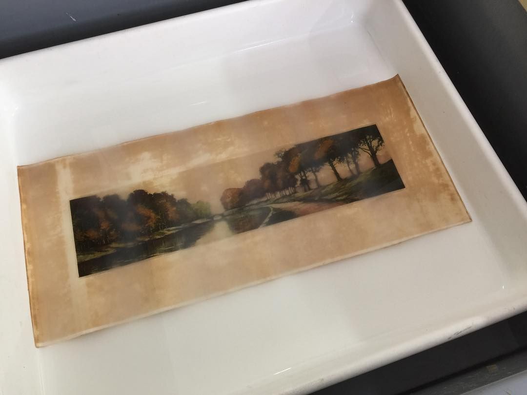 Œuvre de paysage dans un plateau blanc, montrant des arbres, de l'eau et un chemin ; tons bruns et verts.