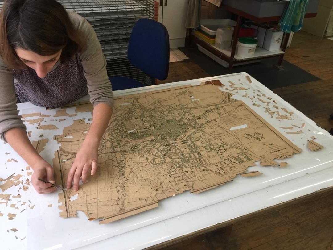 Une femme travaille soigneusement sur une grande carte fragmentée disposée sur une surface blanche, à l'aide de petits outils dans un atelier.