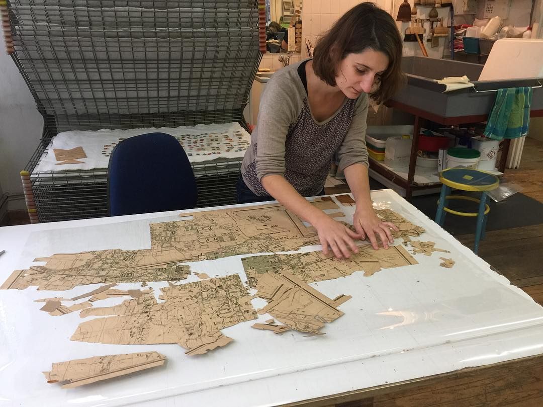 Une femme dispose des morceaux de papier découpé sur une table blanche. Un séchoir et d'autres équipements sont visibles à l'arrière-plan.