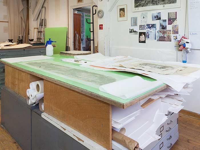 Table de travail en bois dans un atelier, avec des papiers empilés dessus et dessous. Vaporisateur et rouleaux de papier visibles.