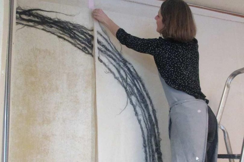 Femme accrochant un panneau d'art en fibre avec des fils noirs sur un mur blanc ; à l'aide d'une échelle.