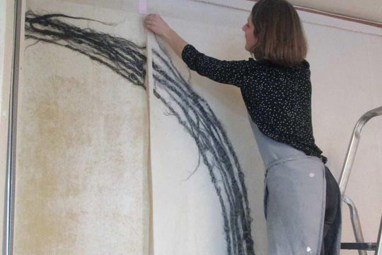Femme accrochant une œuvre d'art textile sur un mur ; l'œuvre présente des lignes noires sur un fond crème et beige.