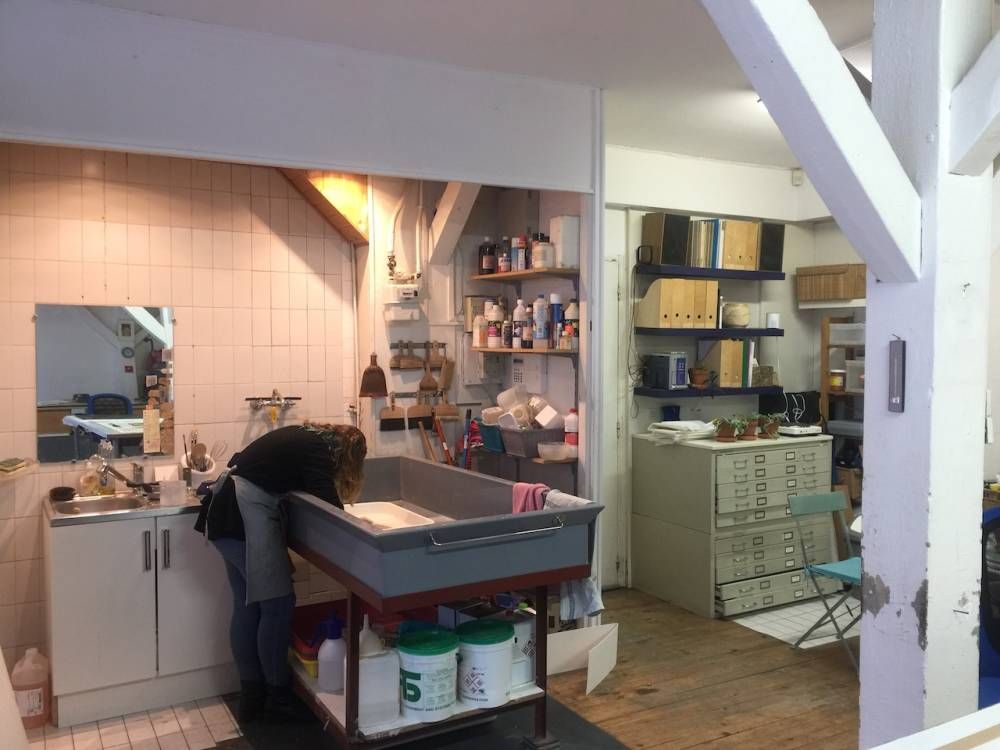 Une personne travaille devant un évier de laboratoire avec un grand plateau. Des étagères remplies de fournitures en arrière-plan. Des poutres en bois et des classeurs.