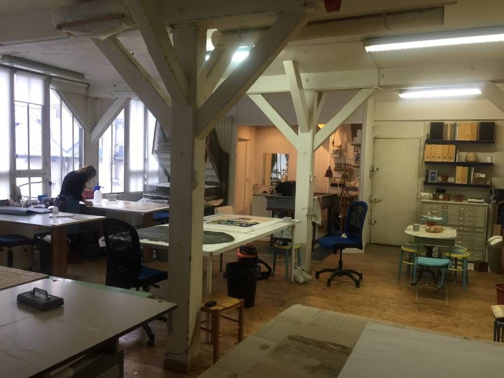Intérieur d'atelier avec poutres en bois blanc, tables et une personne travaillant à une table près d'une fenêtre.