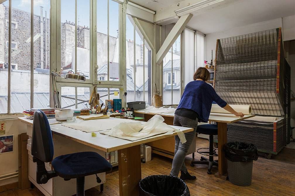 Femme dans un atelier d'art travaillant à un bureau avec de grandes fenêtres donnant sur une ville ; des papiers et des fournitures sont visibles.
