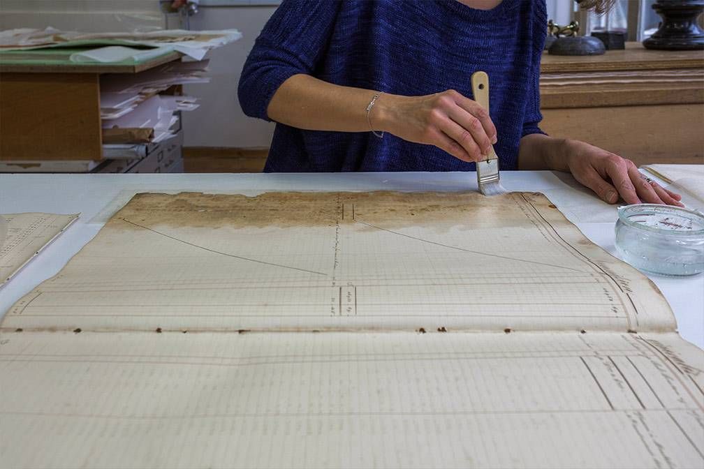 Une personne applique du liquide sur un grand papier vieilli aux marques effacées. Table blanche, pot transparent.