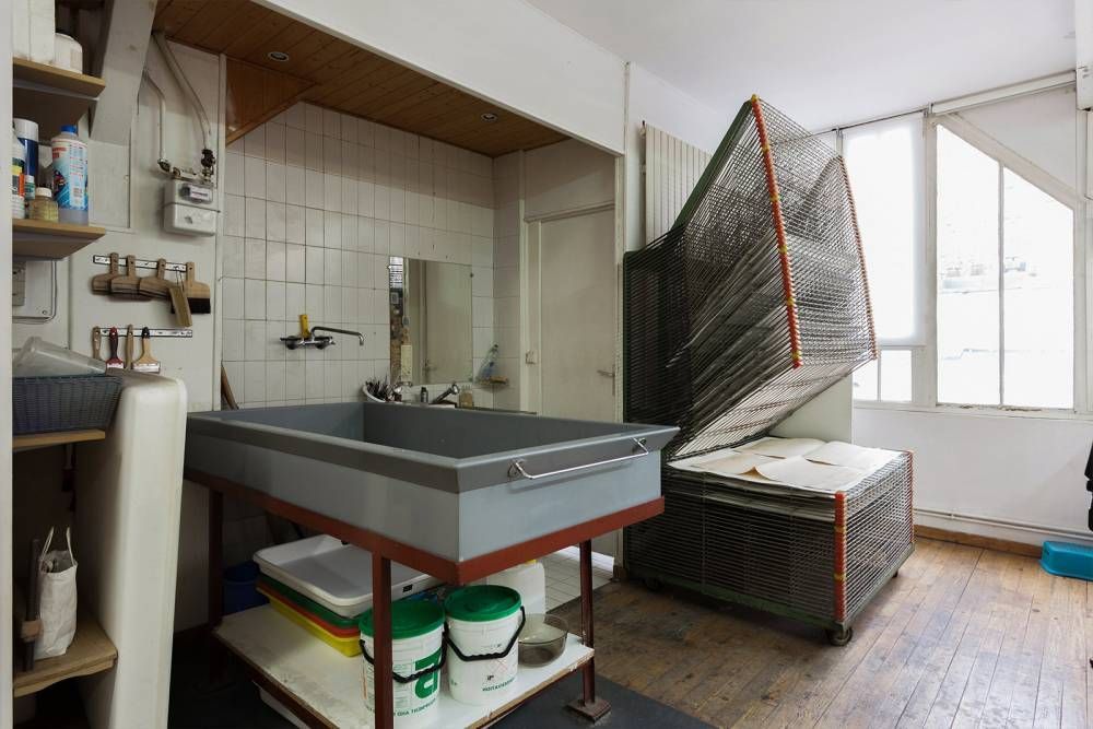 Chambre noire avec plateaux de développement, évier et équipement emballé ; lumière naturelle provenant d'une fenêtre.