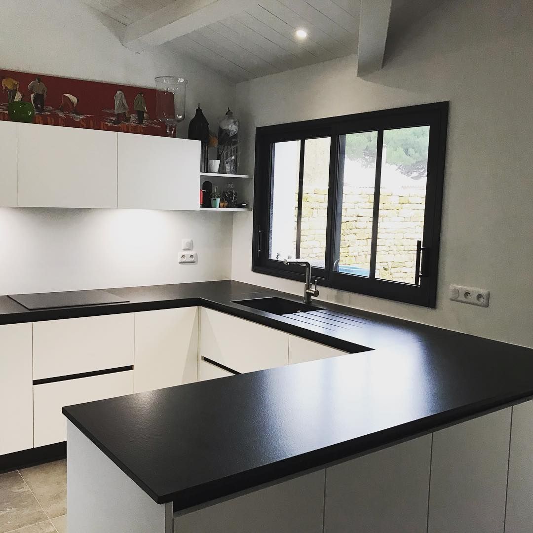 Cuisine moderne avec comptoirs noirs, armoires blanches et une fenêtre à cadre noir laissant entrer la lumière du jour