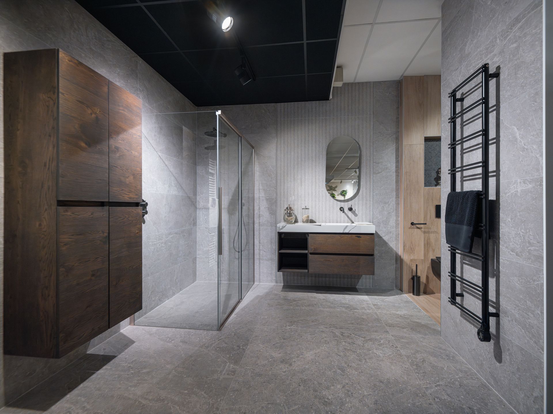 Salle de bains moderne avec meubles foncés, douche en verre, meuble-lavabo, miroir et porte-serviettes dans une pièce carrelée grise