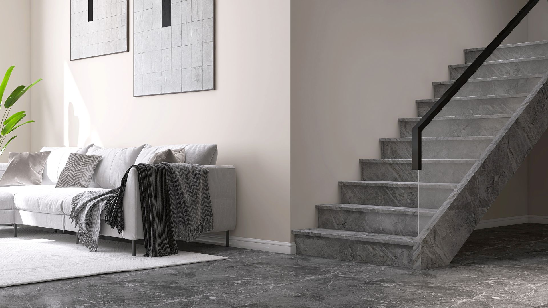 Salon moderne avec un canapé gris, des tableaux encadrés et un escalier sombre sur la droite.
