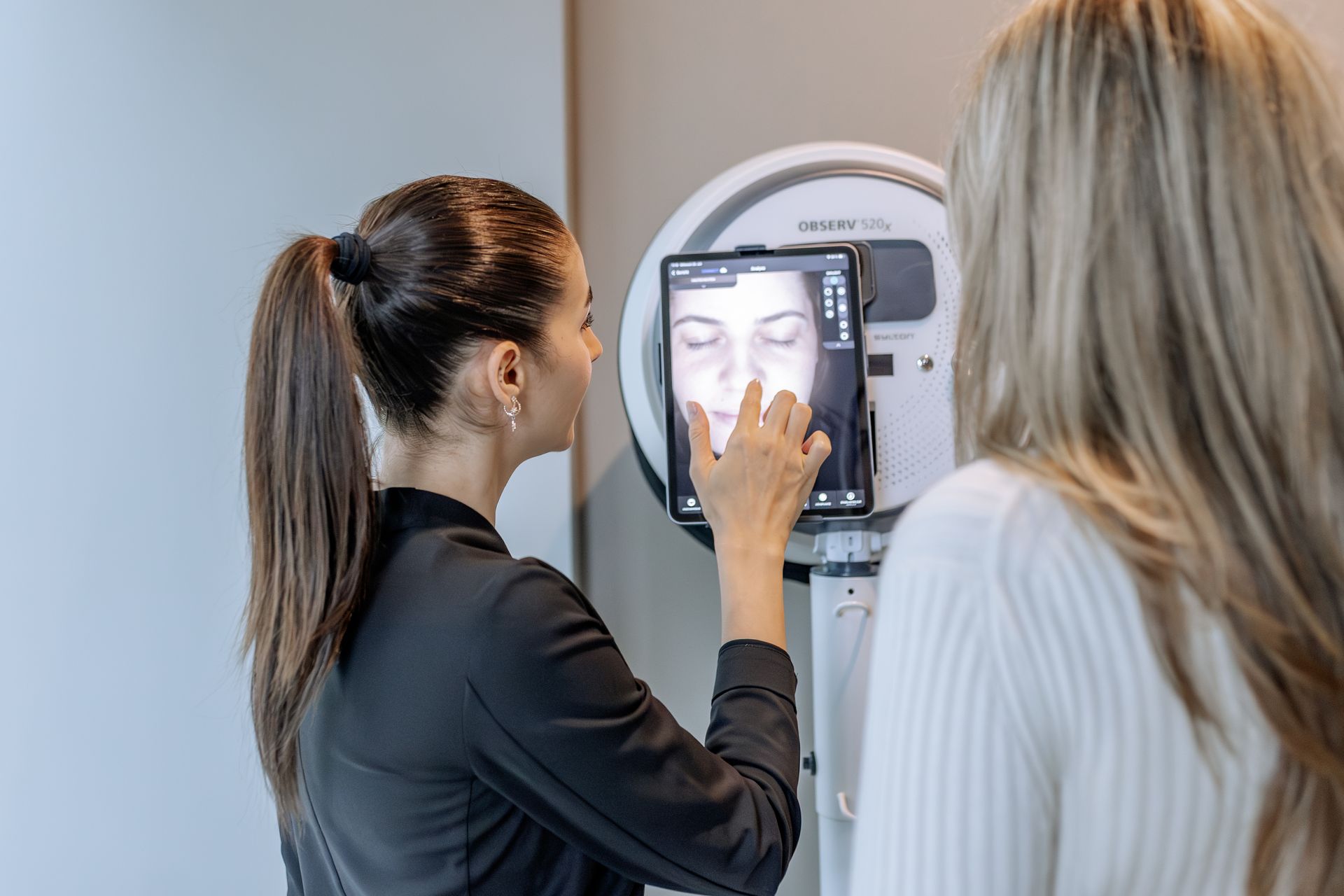 Eine Frau zeigt auf den Bildschirm eines Tablets; eine andere Frau steht neben ihr vor einem Gesichtserkennungsgerät.