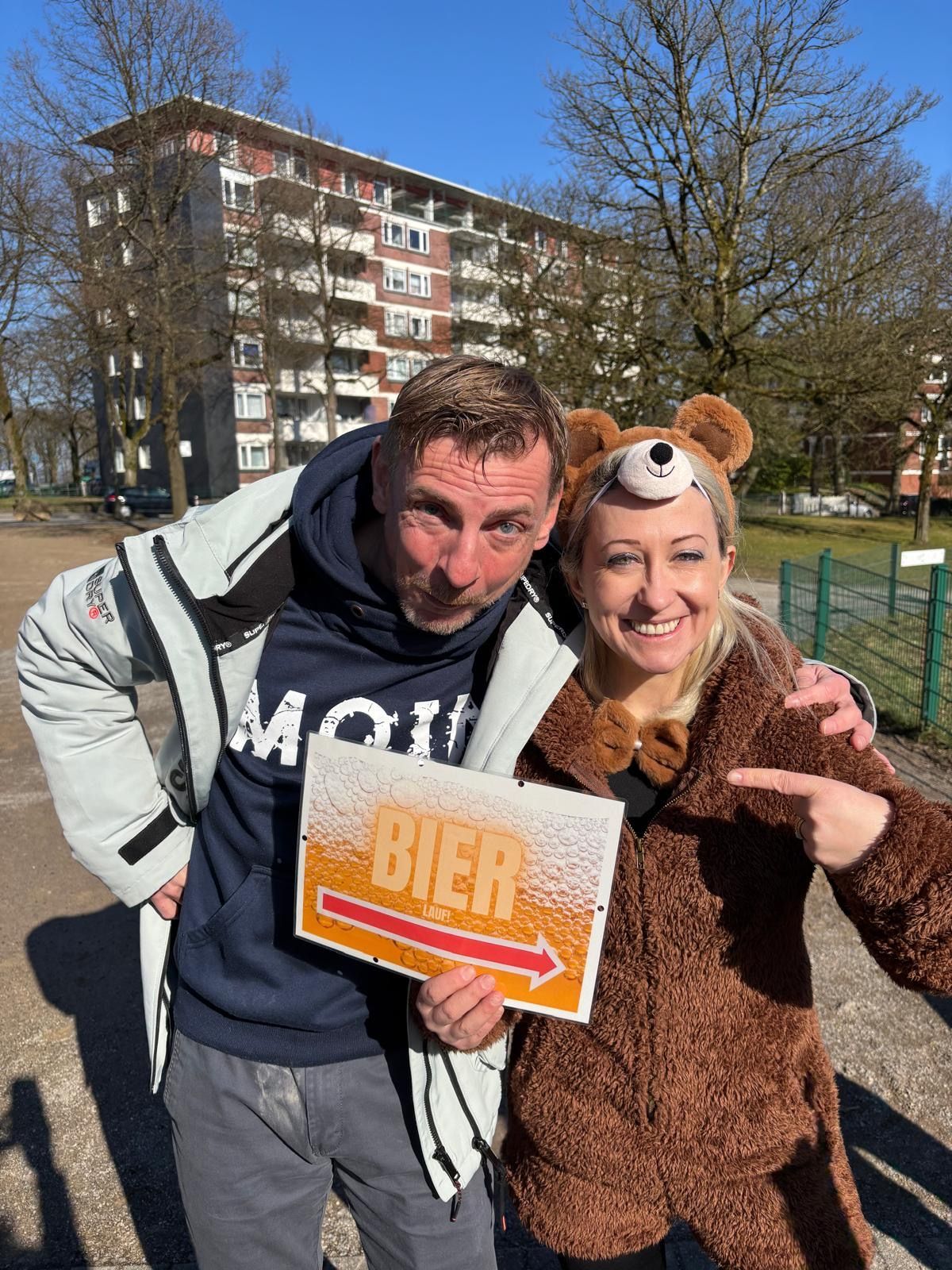 Mann und Frau lächeln, halten ein Bierschild, Frau im Bärenkostüm im Freien.
