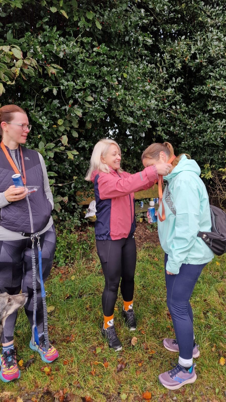 Drei Frauen im Freien; eine richtet die Medaille einer anderen. Grünes Laub im Hintergrund.