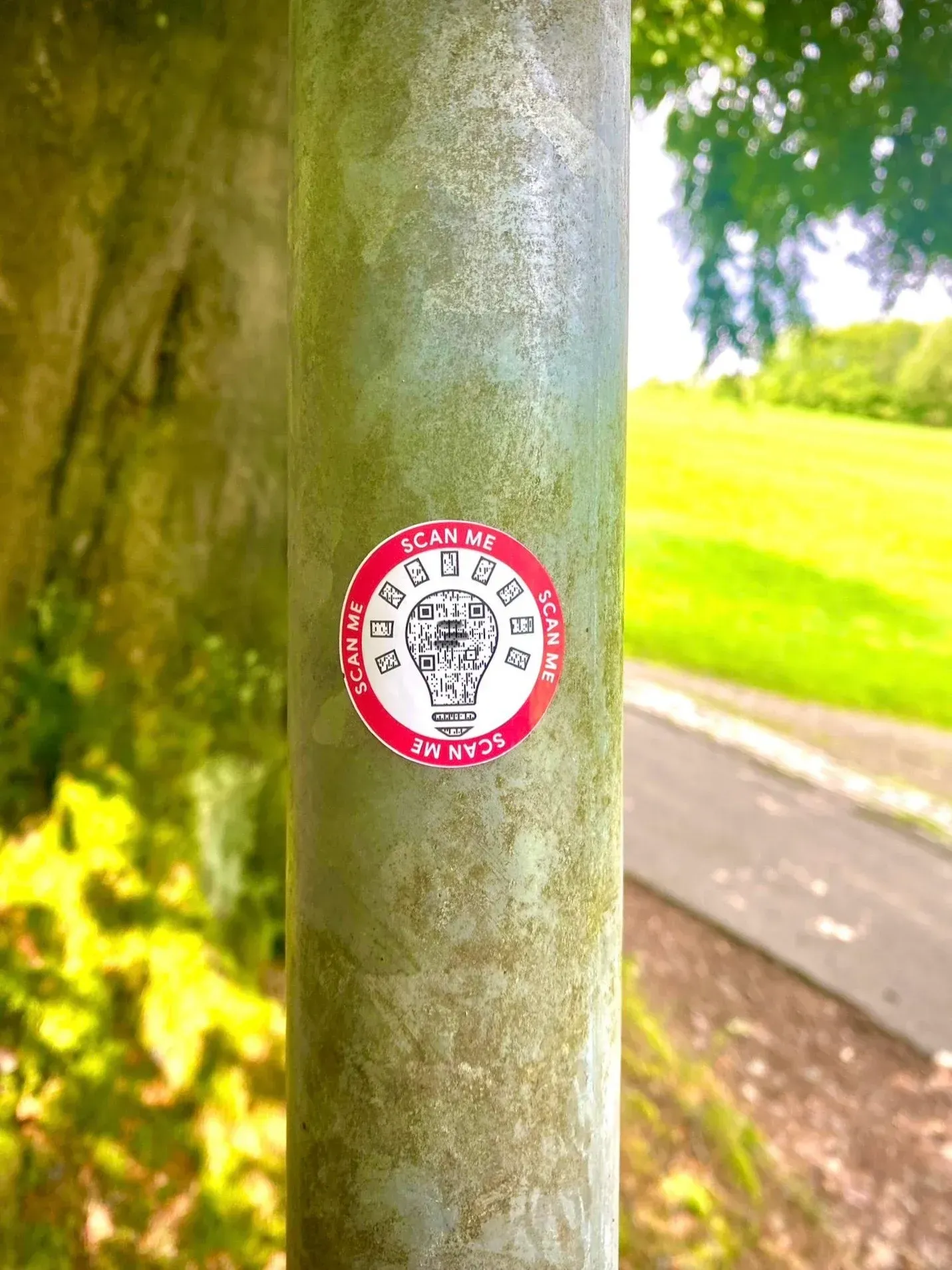 Ein runder Aufkleber mit einer Glühbirnengrafik auf einem Metallpfosten in einer Parkumgebung.