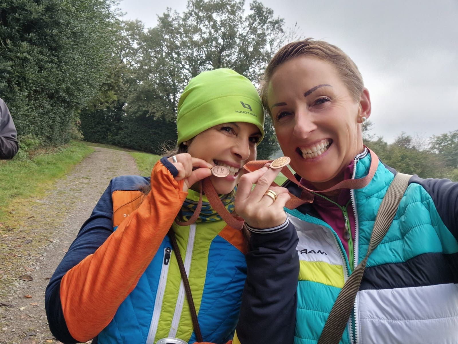 Zwei Frauen lächeln und beißen in Medaillen auf einem Weg im Freien. 