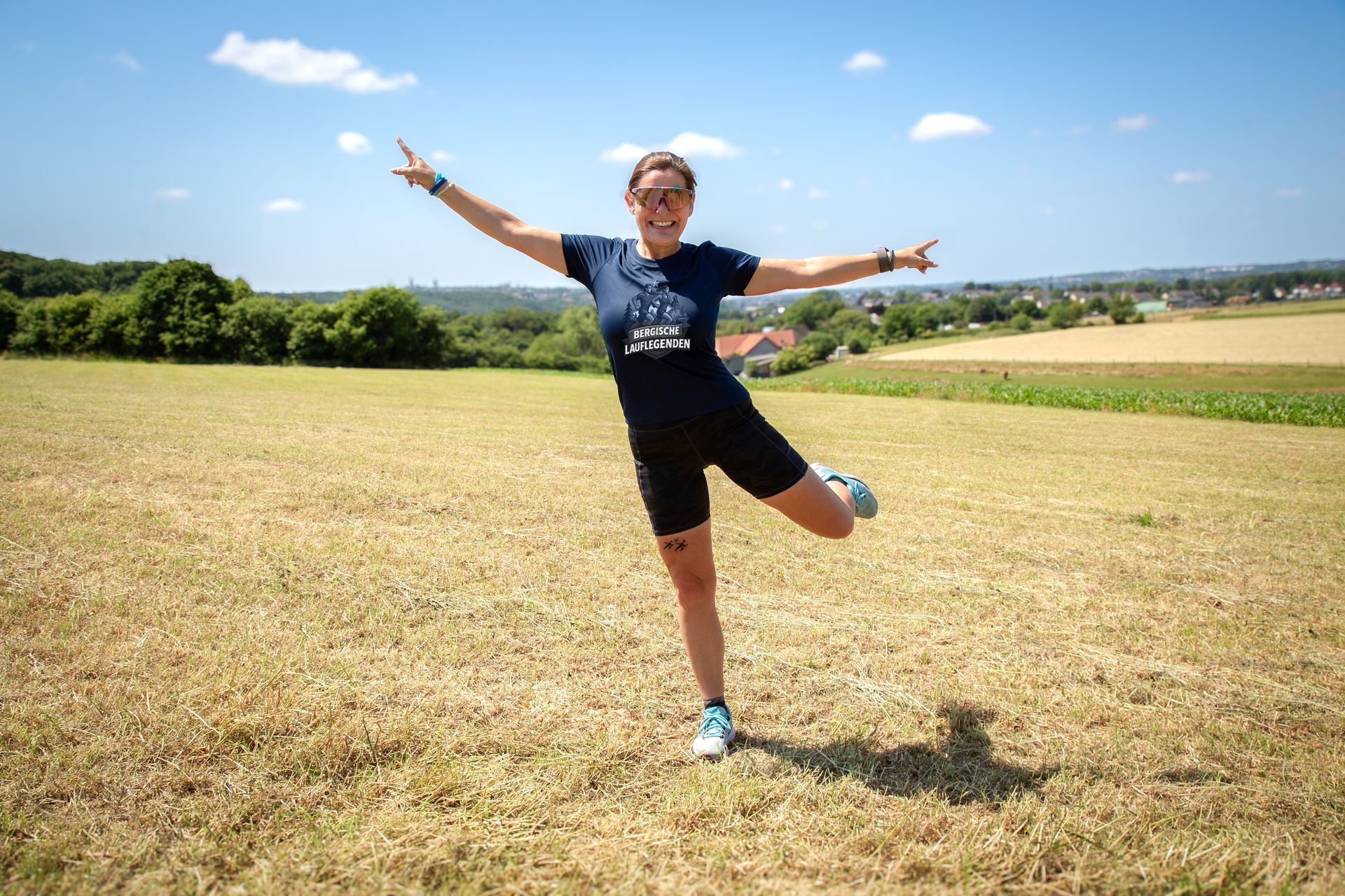 Frau in Sportkleidung, lächelnd, Arme und ein Bein an einem sonnigen Tag auf einem Feld ausgestreckt.