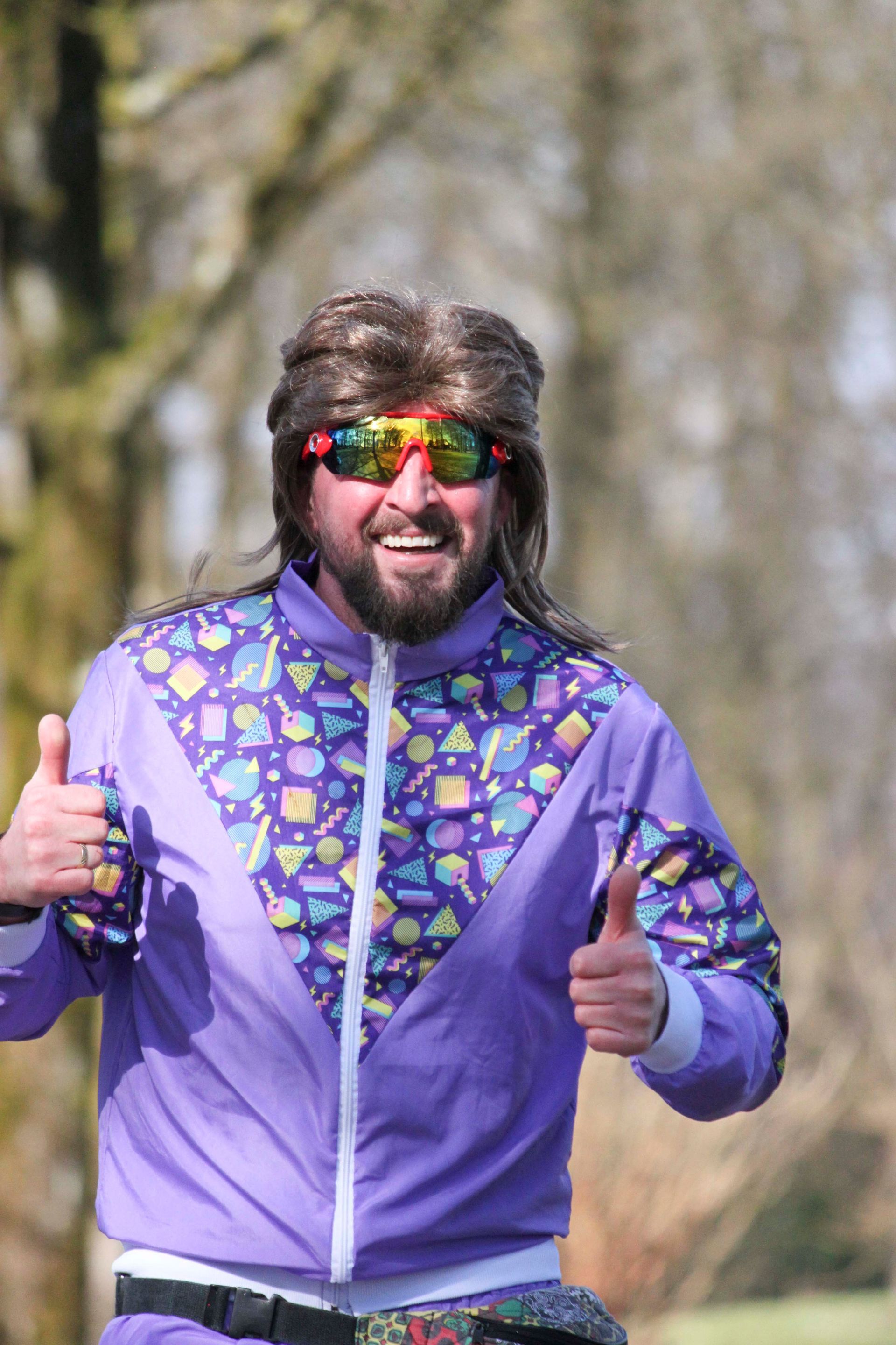 Mann in lilafarbenem Trainingsanzug aus den 80ern, Sonnenbrille und Vokuhila-Perücke zeigt im Freien den Daumen nach oben.