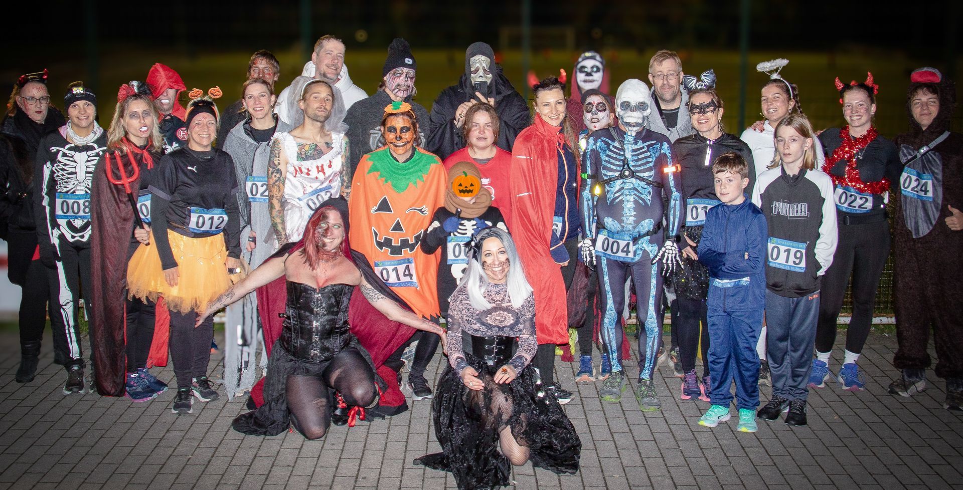 Eine Gruppe von Menschen in Halloween-Kostümen posiert nachts im Freien, mit Kürbissen und gruseligen Outfits.