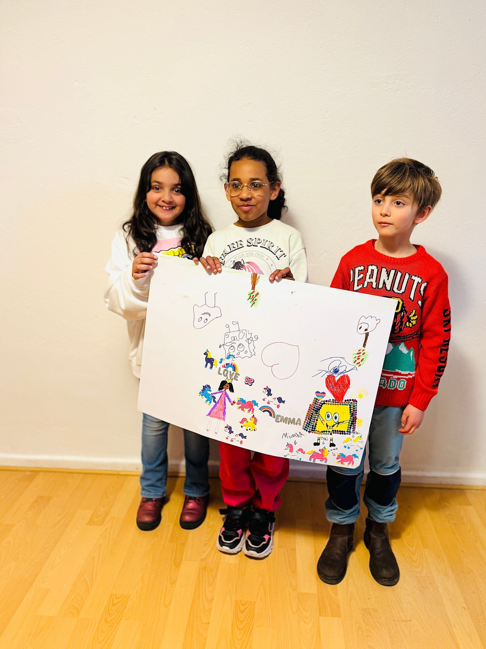 Drei Kinder stehen in einem Zimmer und halten ein großes, handgefertigtes weißes Poster mit bunten Zeichnungen in den Händen.