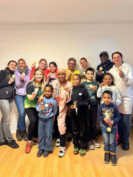 Eine Gruppe von Menschen unterschiedlichen Alters steht in einer Reihe in einem Innenraum, viele halten kleine Goldmedaillen in den Händen und lächeln in die Kamera.