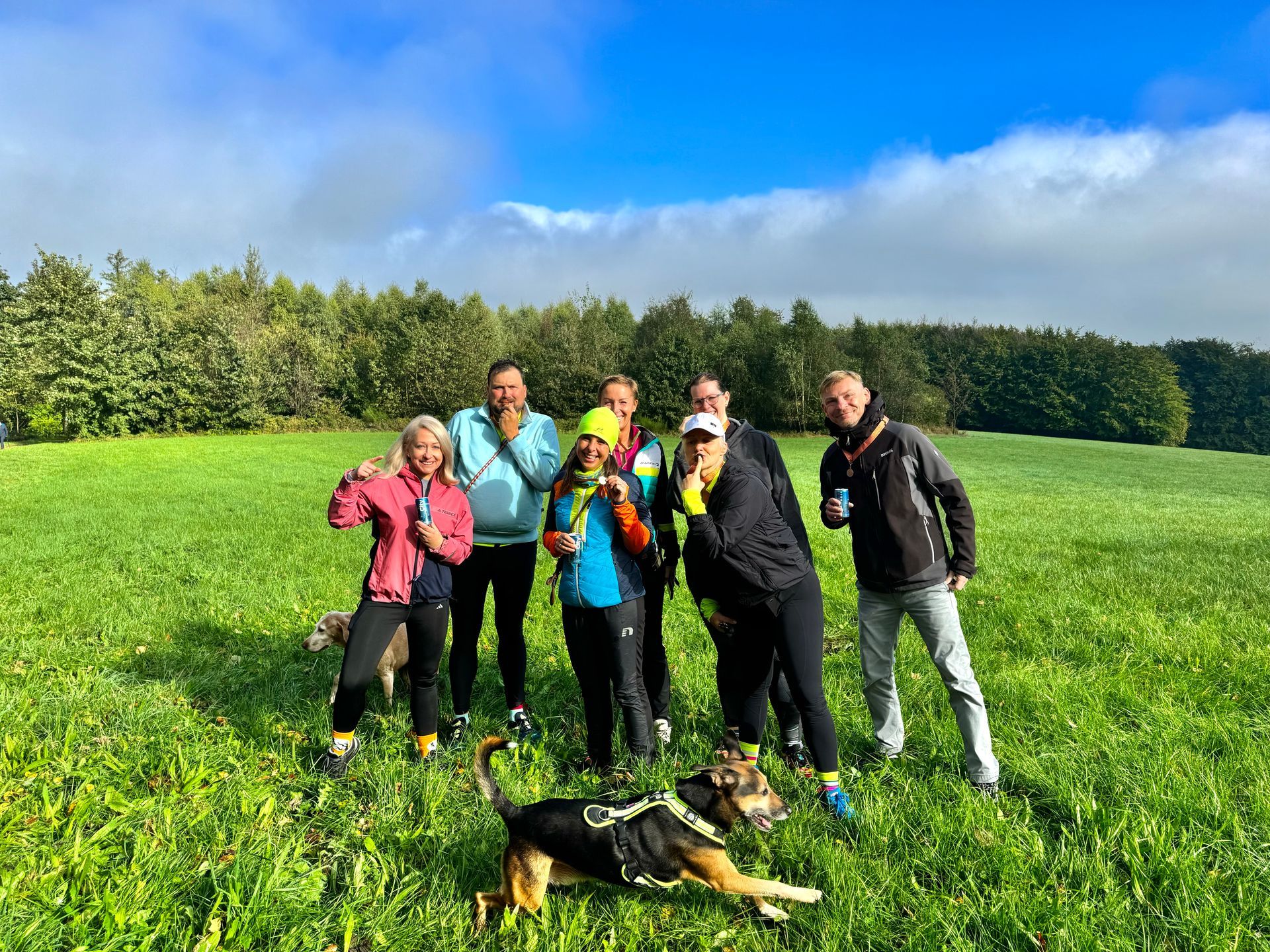 Gruppe von Menschen mit einem Hund auf einer Wiese mit Bäumen unter blauem Himmel.