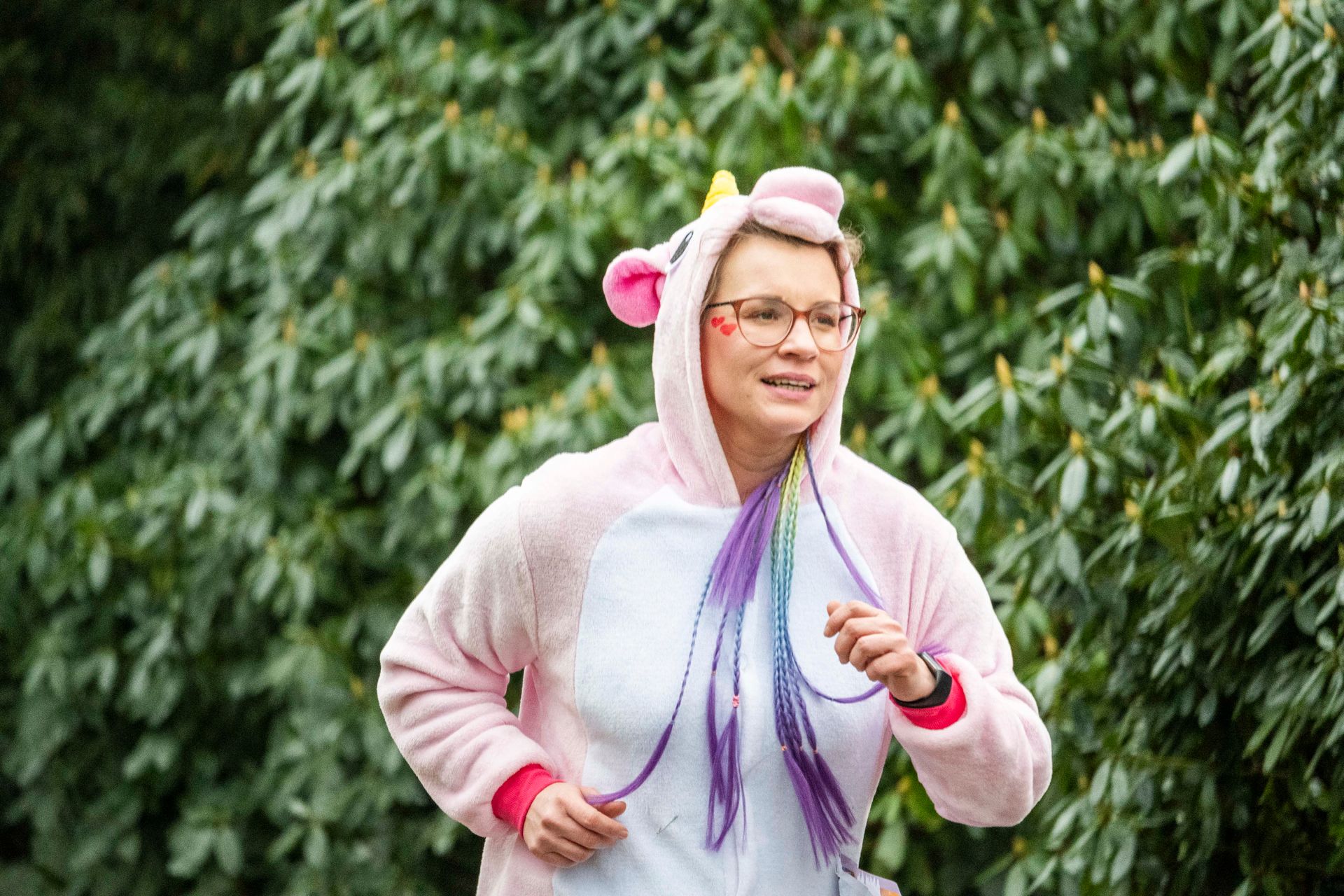 Frau rennt in einem rosa Einhorn-Onesie und mit Brille vor grünem Laub.