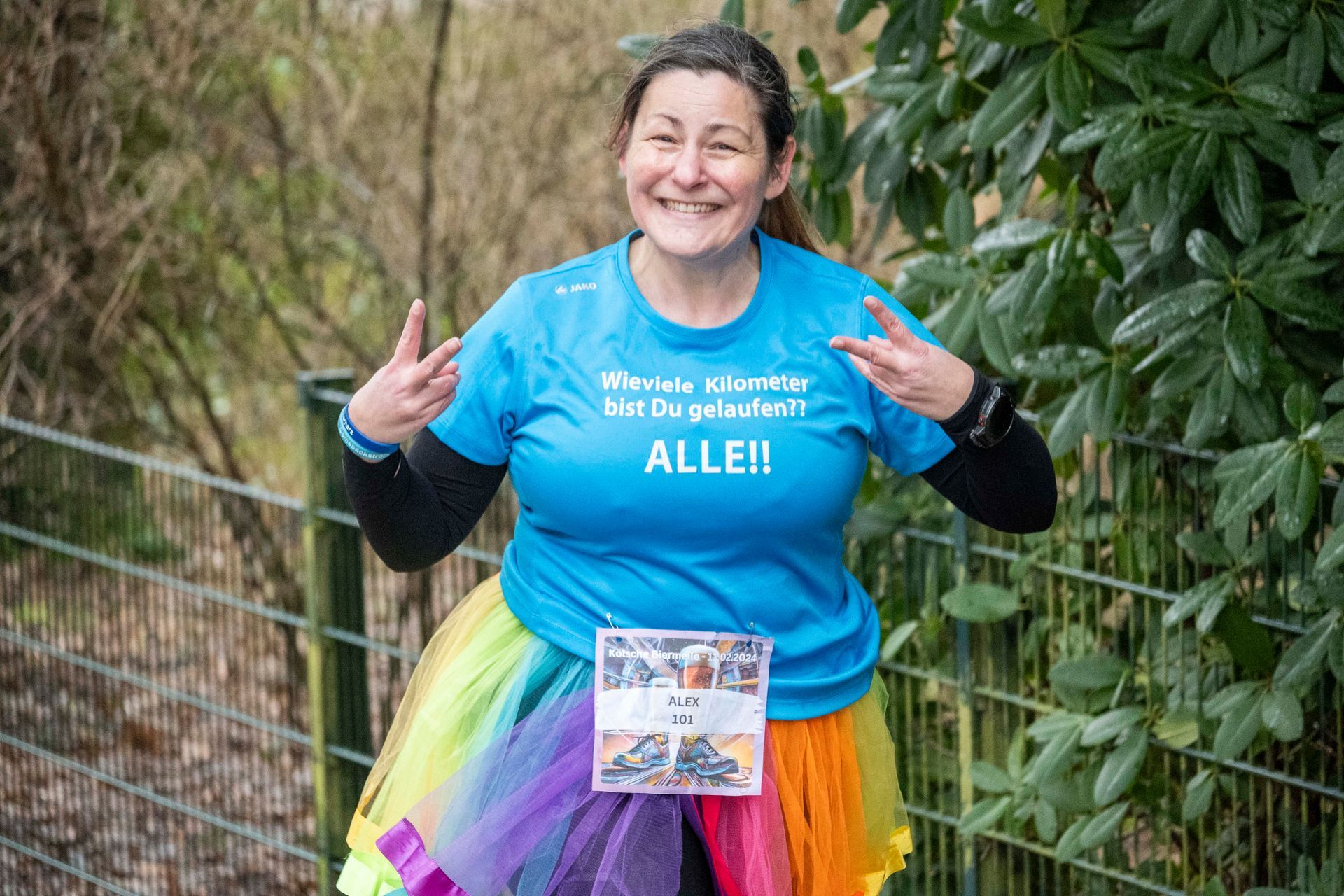 Frau im Regenbogen-Tutu macht während eines Rennens Peace-Zeichen; auf dem blauen Shirt steht „ALLEI!“ auf Deutsch.