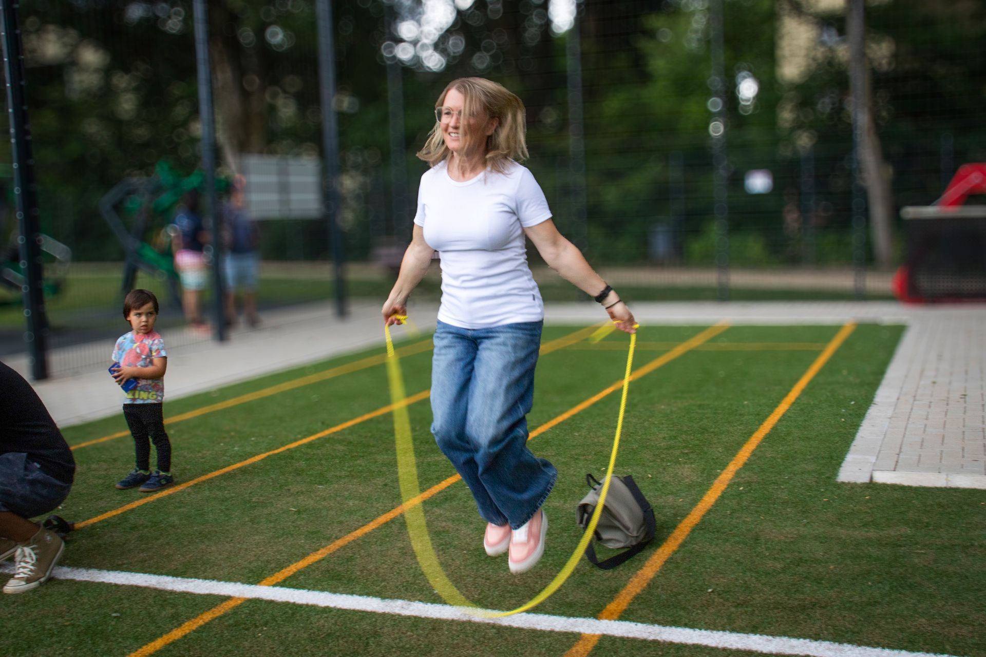 Eine Person springt auf einem Sportplatz Seil; im Hintergrund sind ein Kind und ein Trainer zu sehen.