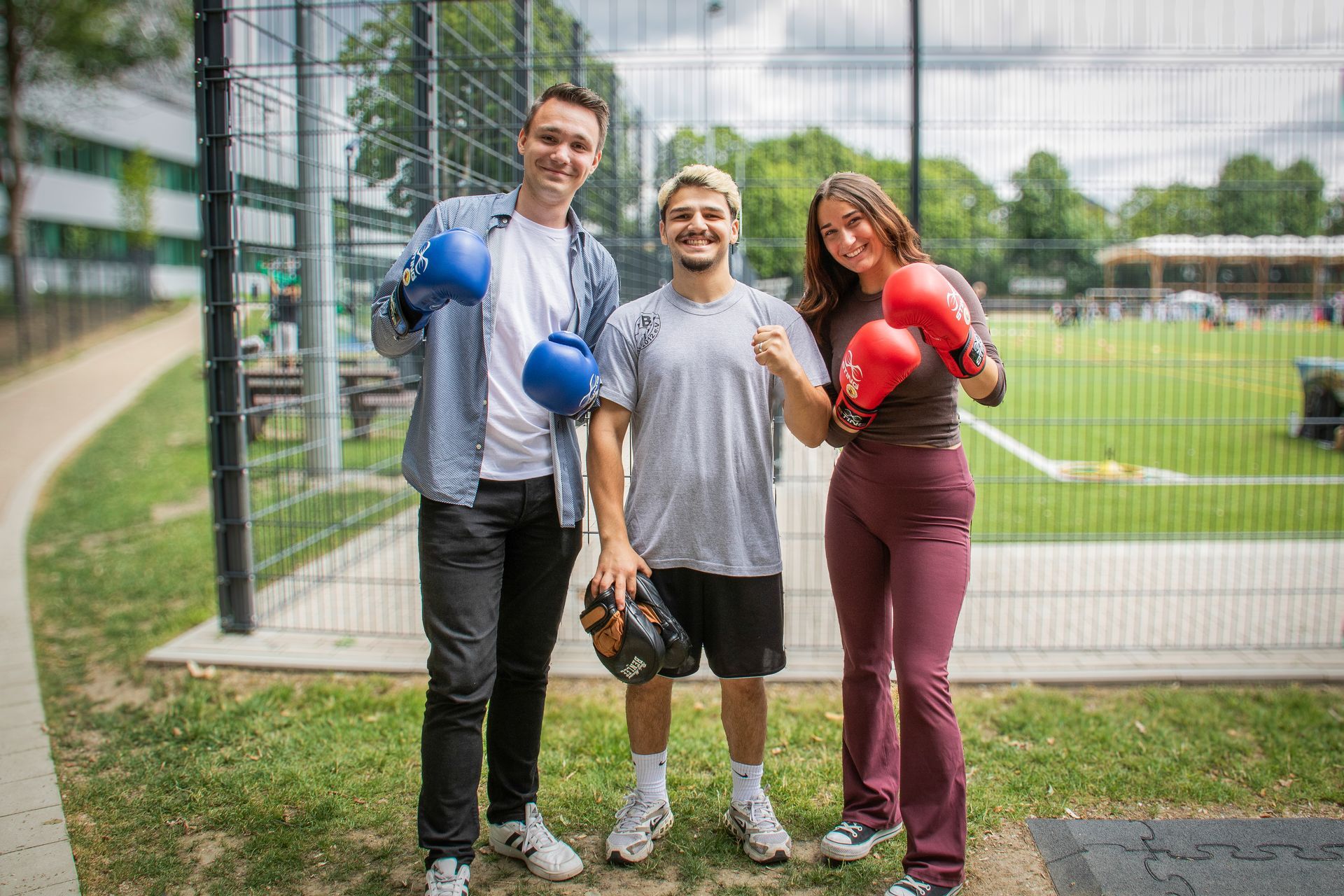 Drei Personen mit Boxhandschuhen posieren lächelnd vor einem umzäunten Sportplatz.