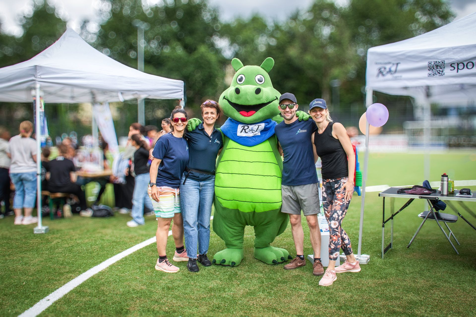 Menschen posieren mit einem grünen Maskottchen auf einem Sportplatz bei einer Freiluftveranstaltung