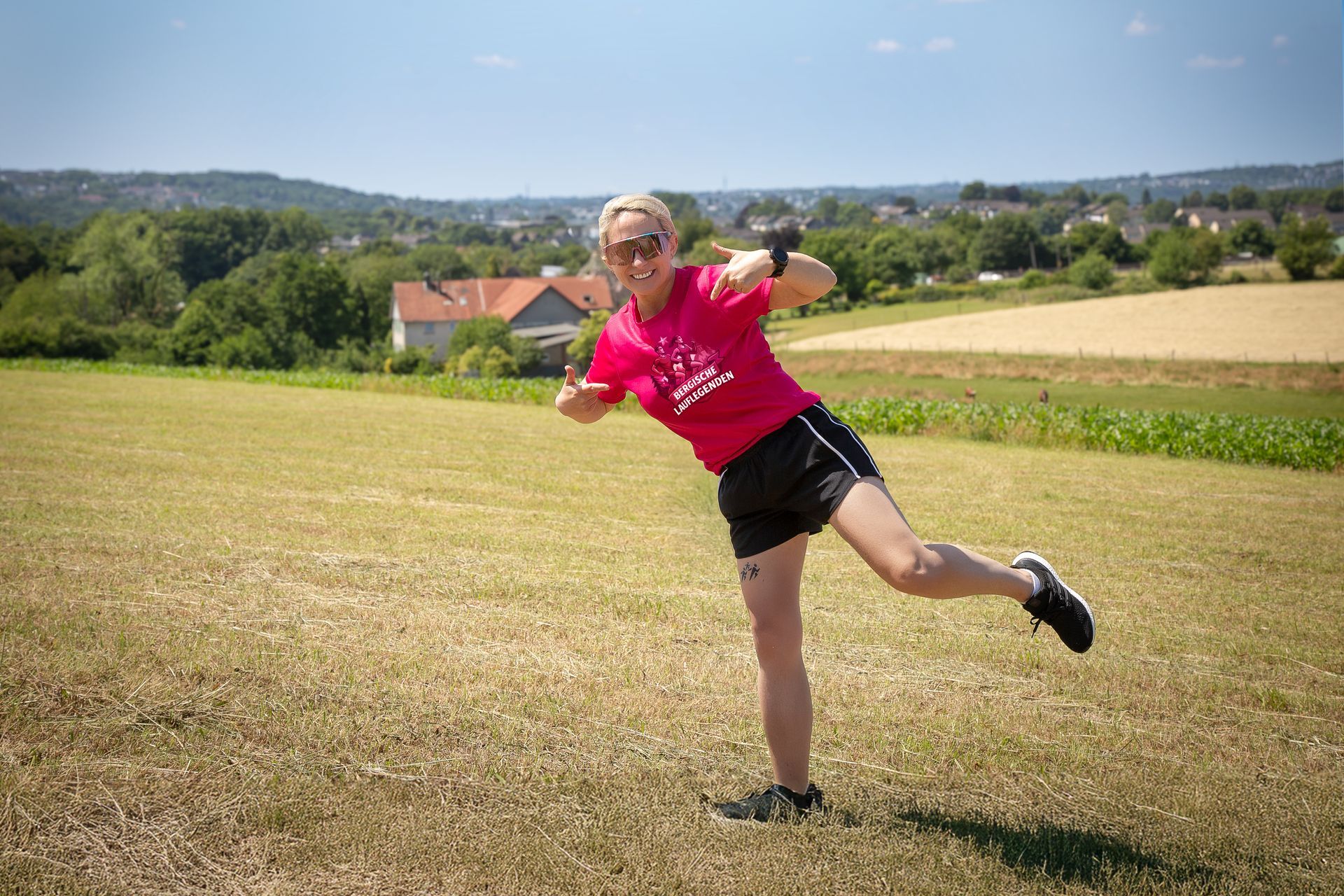 Frau in rosa Hemd legt Bein auf Feld, grüne und braune Landschaft im Hintergrund.