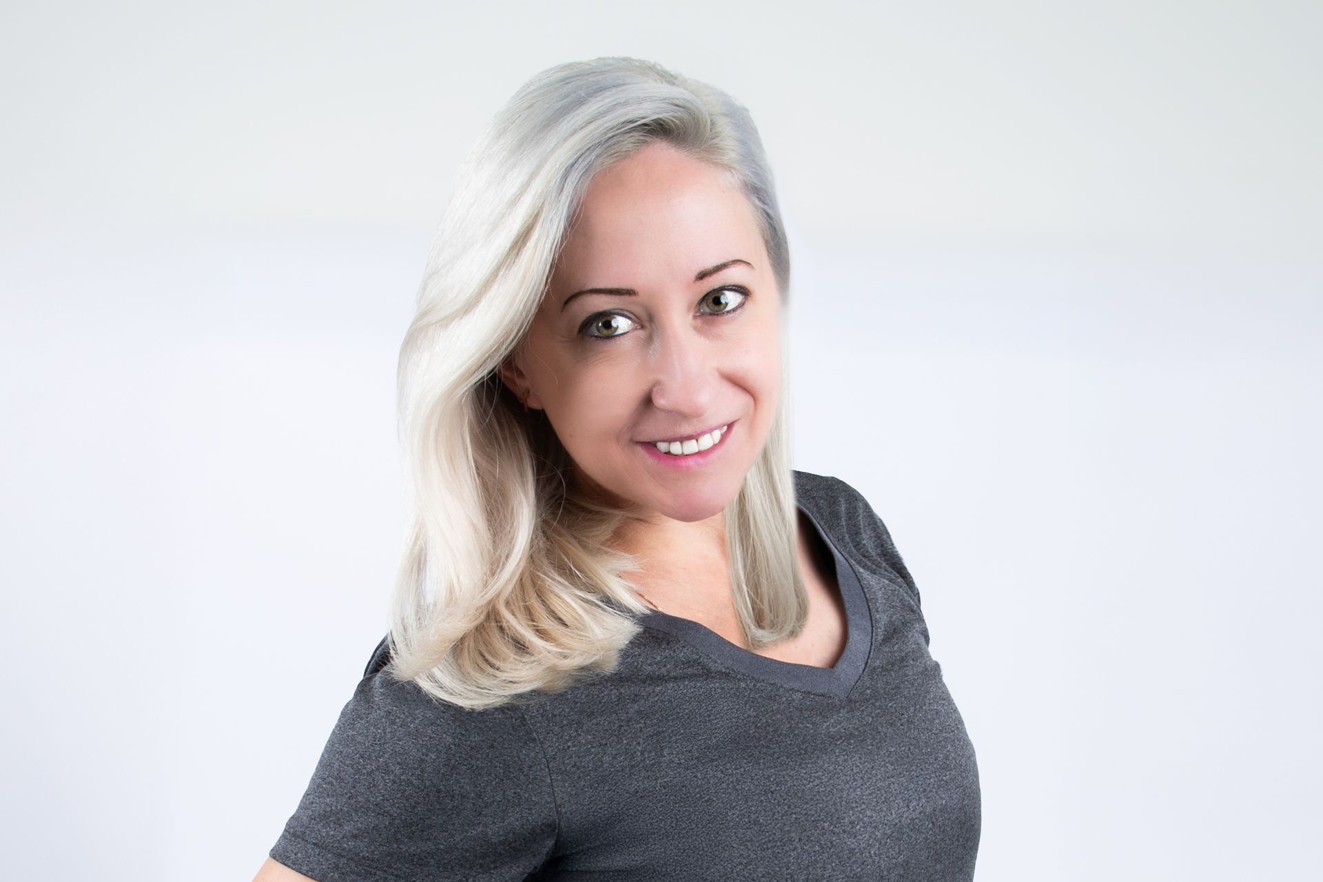 Frau mit grauem Haar, leicht lächelnd, trägt ein graues Hemd vor weißem Hintergrund.