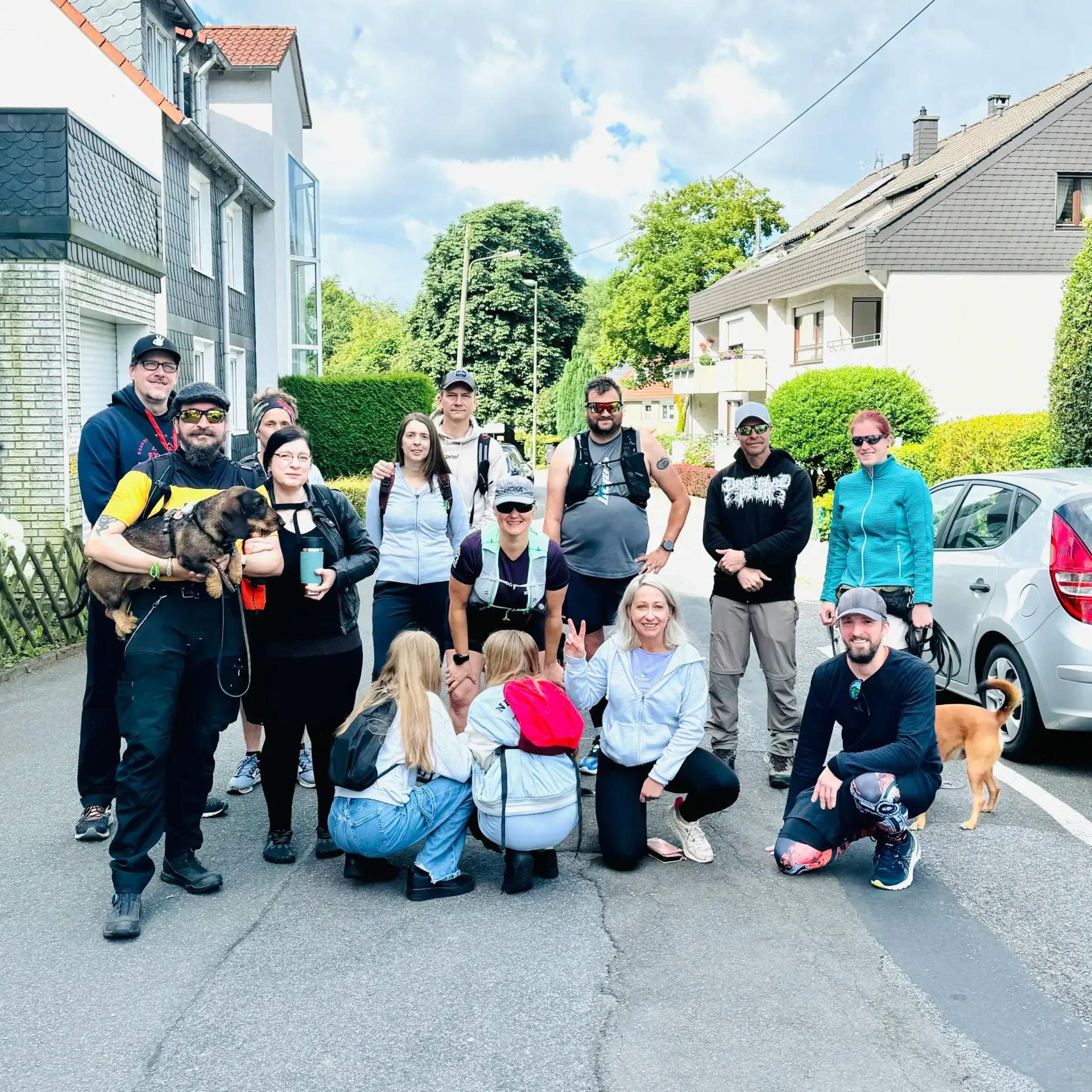 Eine Gruppe von Menschen posiert im Freien auf einer Straße. Einige halten einen Hund, andere tragen Hüte oder Rucksäcke.