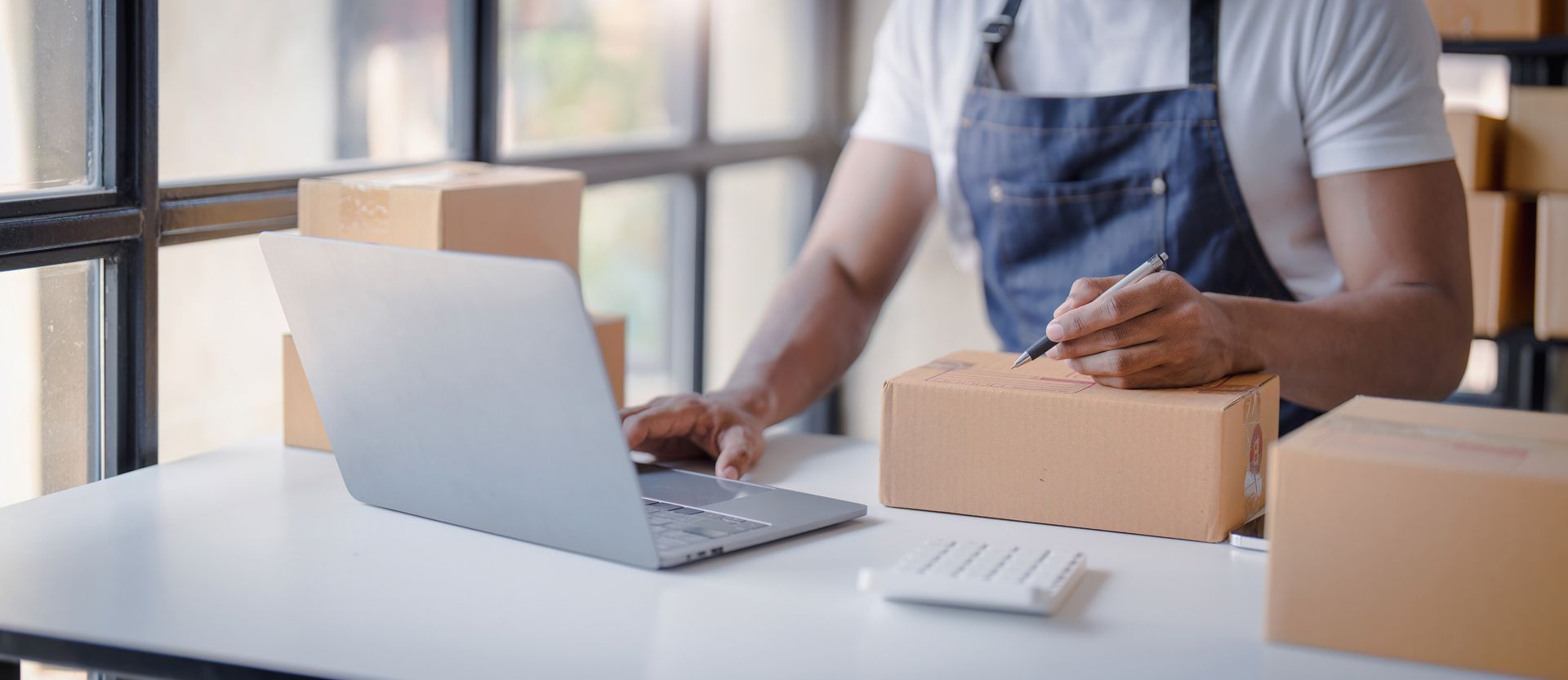 Persona con delantal, escribiendo en un paquete mientras trabaja en una computadora portátil en una mesa con cajas de cartón.