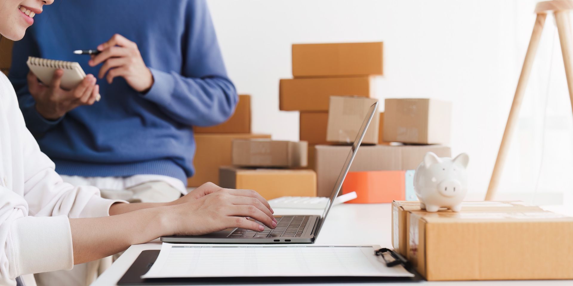 Dos personas trabajan en un escritorio con una laptop, cajas y una alcancía. Una escribe mientras la otra toma notas.