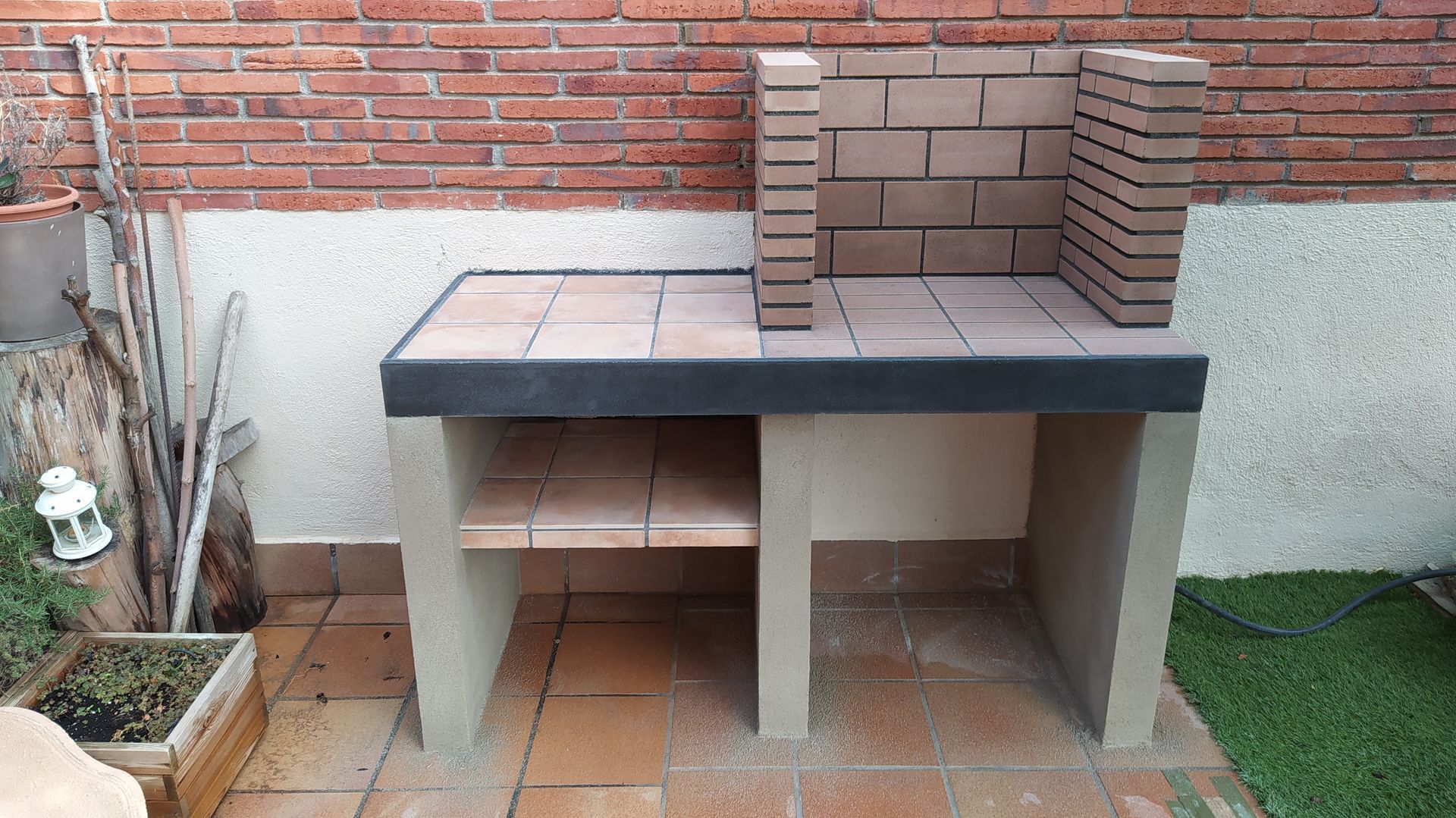 Una parrilla de ladrillos está colocada en un patio de baldosas al lado de una pared de ladrillos.