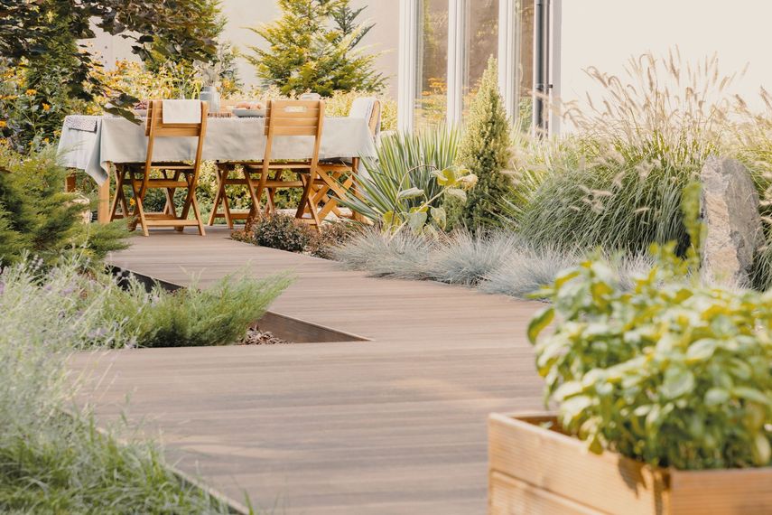 Ensemble de salle à manger en bois pour terrasse, bordant une allée de jardin sinueuse ornée de hautes herbes et de plantes en pot.