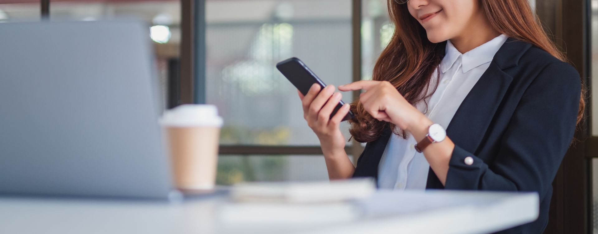 Une femme en blazer bleu marine utilise son smartphone à la table d'un café, avec une tasse de café et un ordinateur portable en arrière-plan.