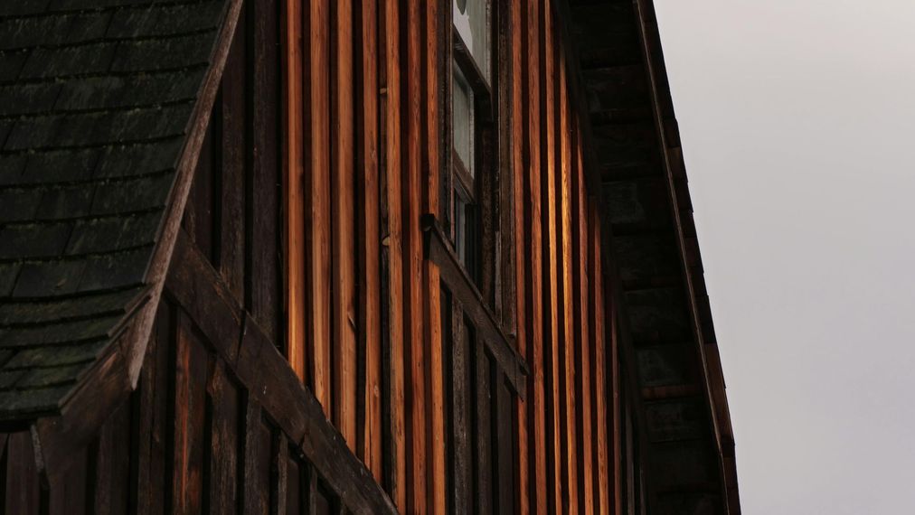 Gros plan sur la façade d'un bâtiment en bois sombre, avec un bardage vertical orange-brun et un toit en bardeaux.